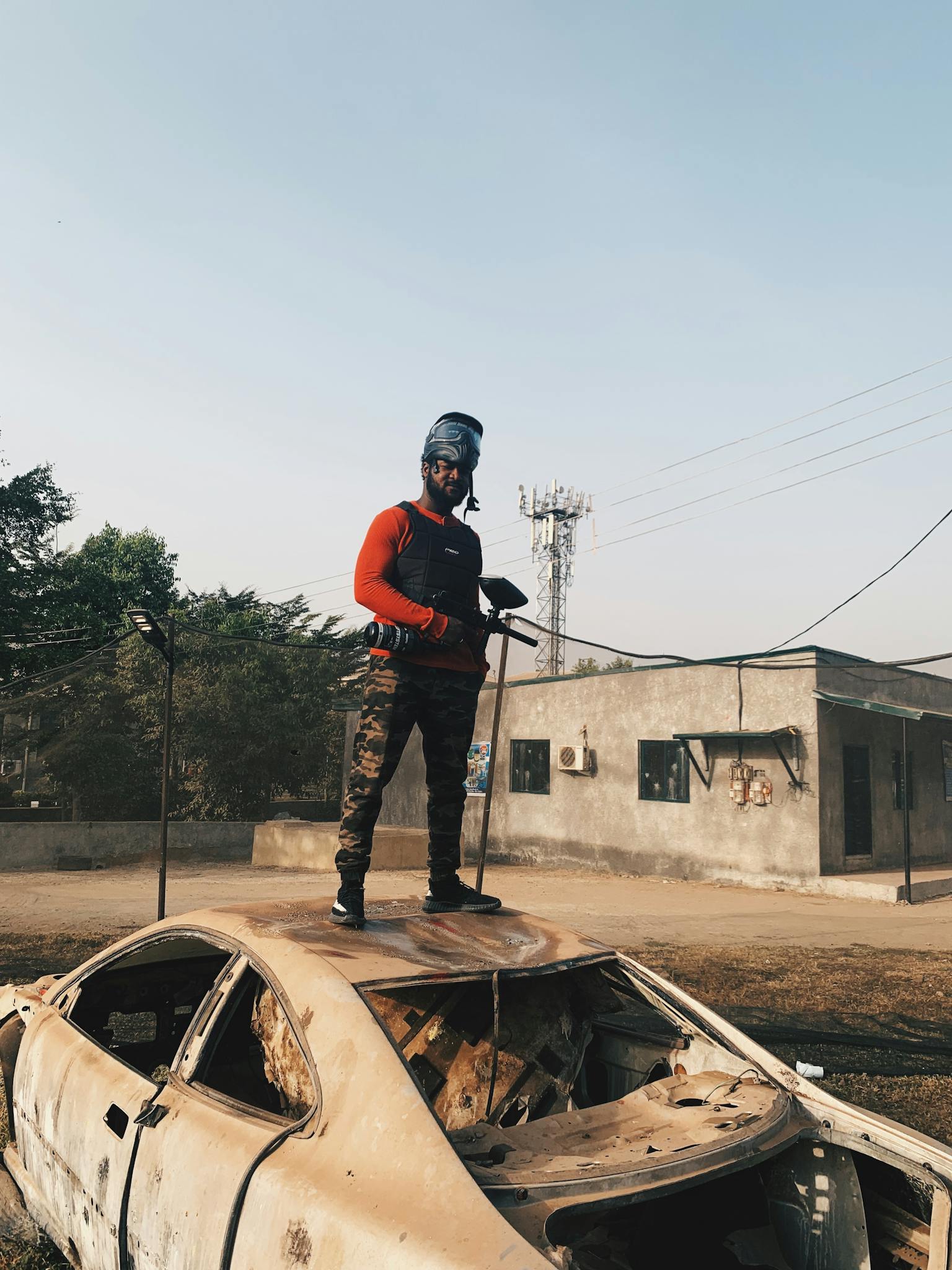 A man in paintball gear stands on a wrecked car in an outdoor setting, ready for action.