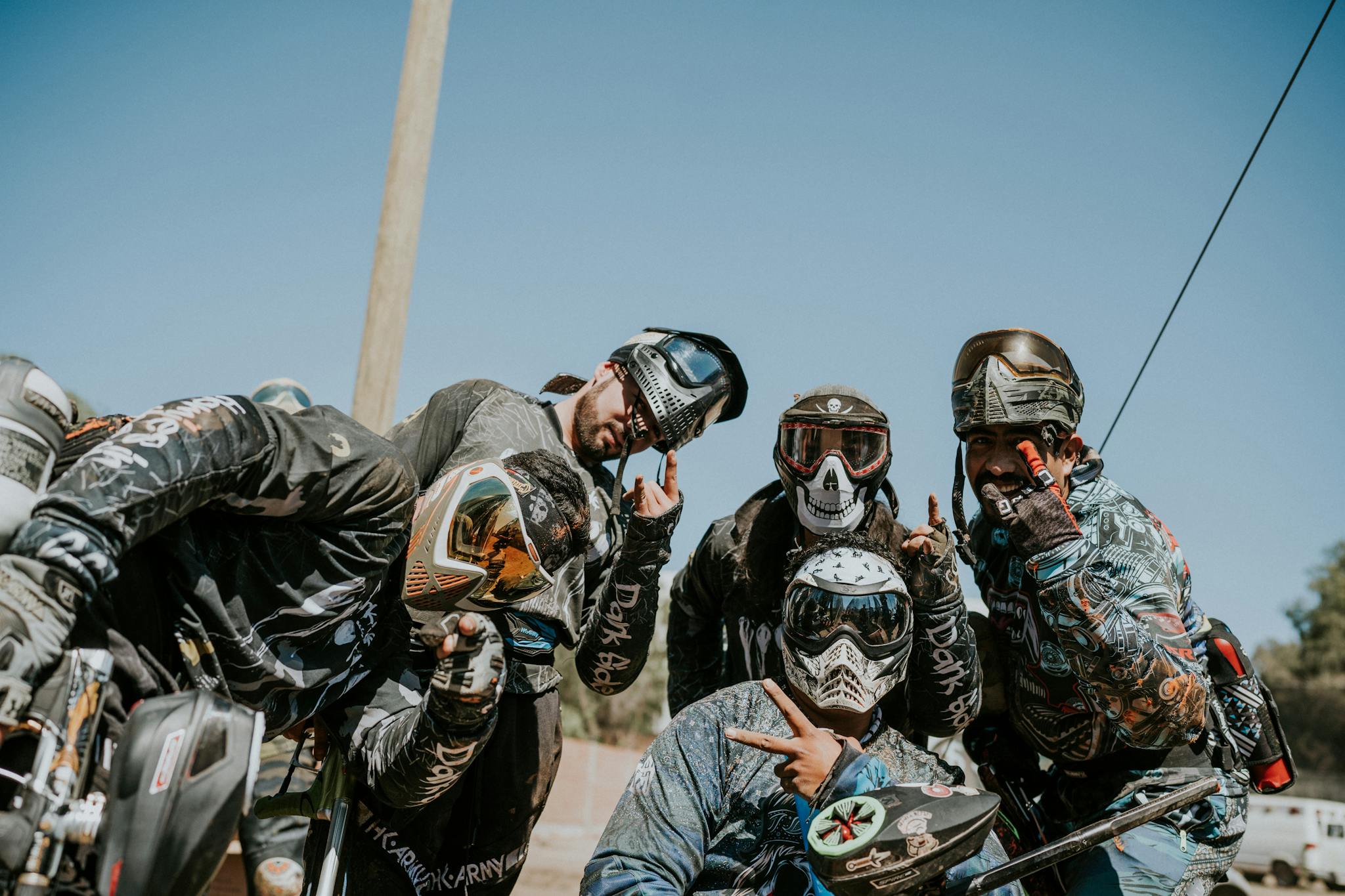 Five paintball players in gear, posing outdoors with hand gestures and goggles.
