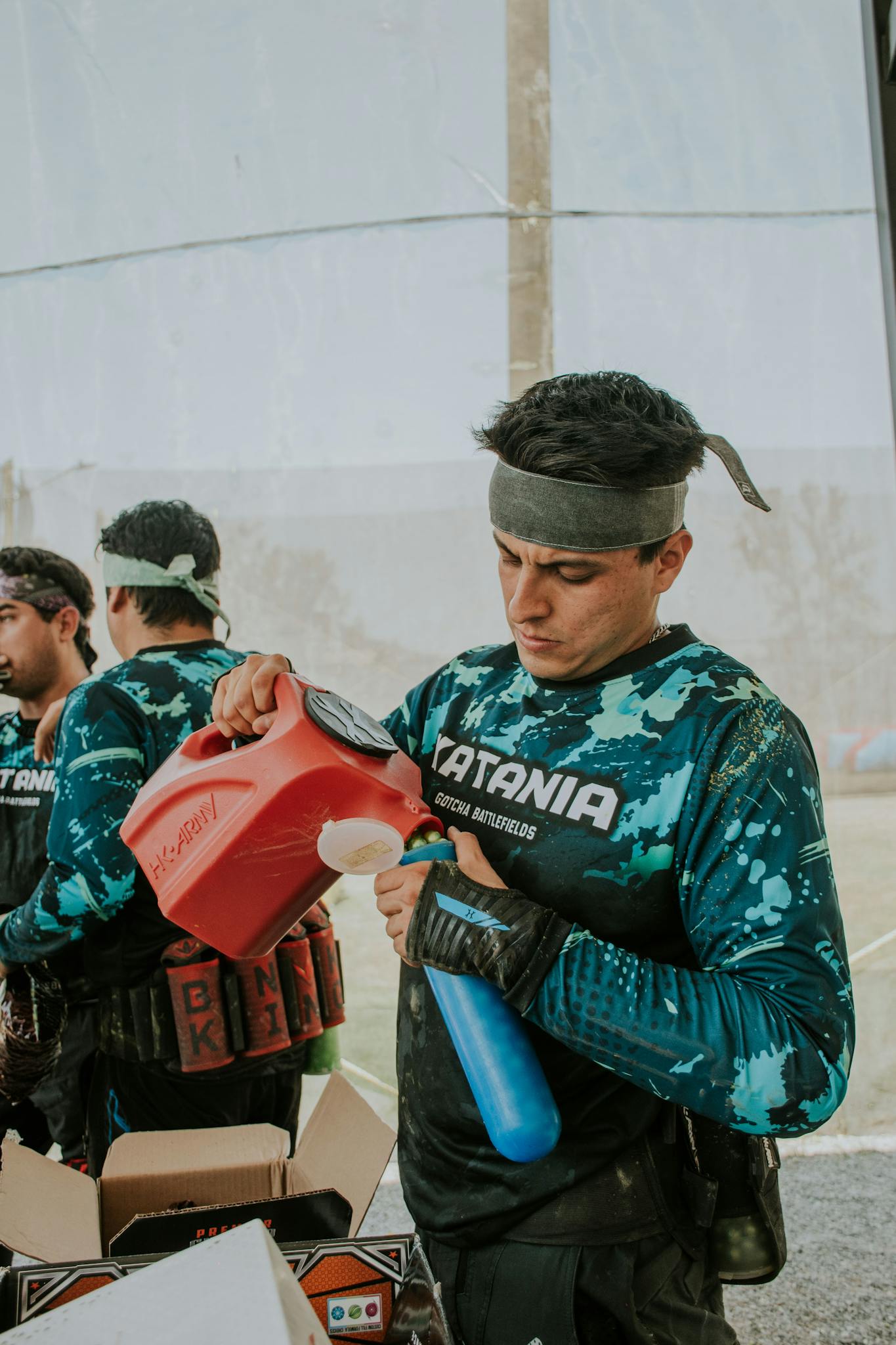 Young man refilling paintball marker tank at outdoor field.